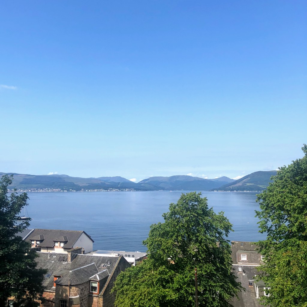 View across the Firth of Clyde from Gourock: nearly-cloudless skies above the hills and the river, with roofs and trees in hte foreground. 
