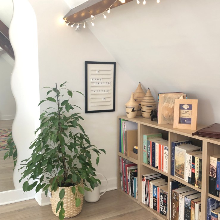 Corner of a room with low-height bookshelves, a floor-standing plant, fairy lights strung from a beam, and a letterboard with 'Frustrated Nester' spelled out.